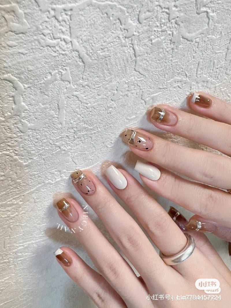 mau-nail-don-gian-cardina__153__9c23dde1a8764730b190cb6bedbe7014 Hai bàn tay với móng được sơn trang trí tinh tế, gồm màu trắng và nâu, nền là tường có kết cấu thô.