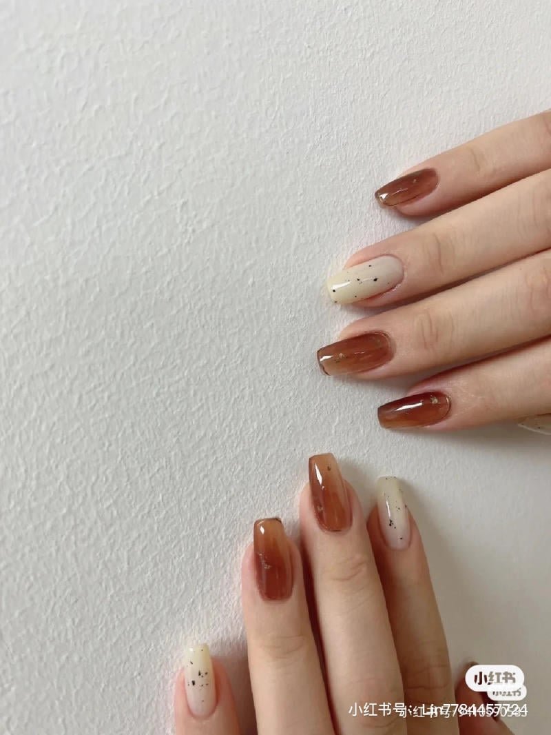 mau-nail-don-gian-cardina__158__9c330bdbf9d74143b19404ef12d2866c Hai bàn tay với móng tay được sơn màu nâu và trắng, có họa tiết chấm đen, trên nền tường trắng.