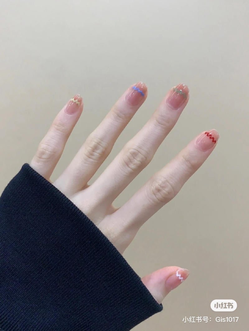mau-nail-don-gian-cardina__3__8c7330d8d3a140928daba4a8c555f39f Một bàn tay với móng tay được sơn màu sắc khác nhau, mỗi móng có họa tiết đường zigzag, áo tay dài màu đen.