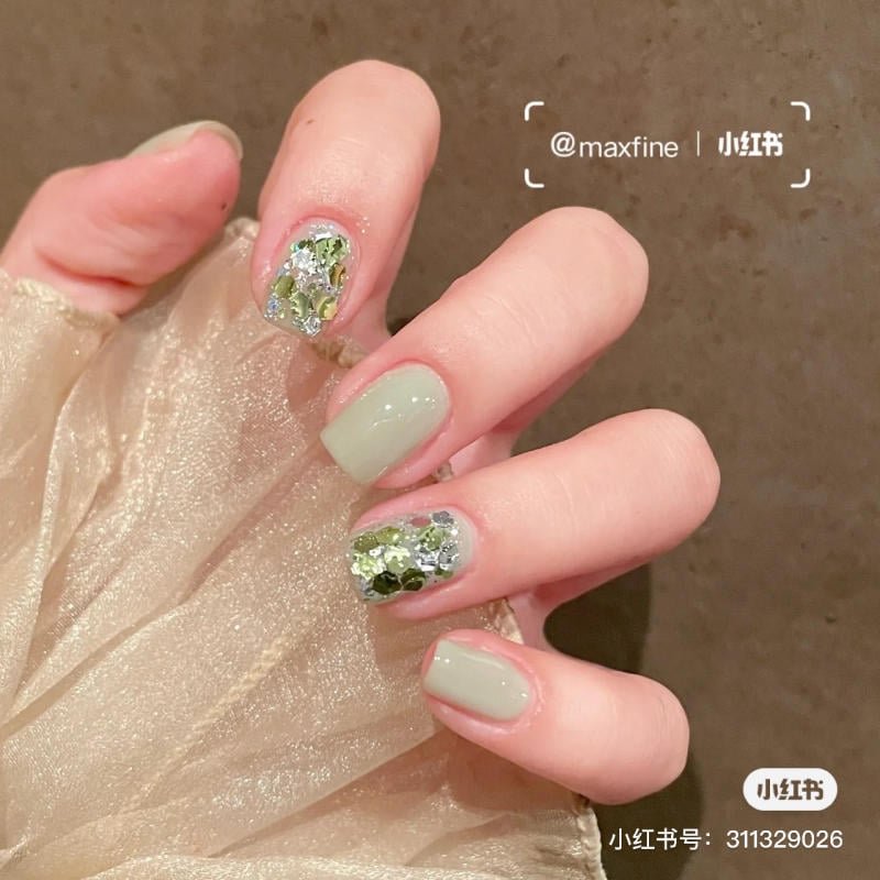Bàn tay với móng tay sơn màu xanh, hai ngón có đính kim tuyến lấp lánh, nền vải voan màu be.